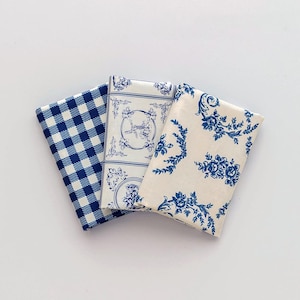 Könnte beinhalten: Drei gefaltete Stoffservietten mit verschiedenen Mustern. Eine ist blau und weiß kariert, eine andere hat einen weißen Hintergrund mit blauen Blumenmustern, und die dritte hat einen weißen Hintergrund mit blauen Illustrationen.