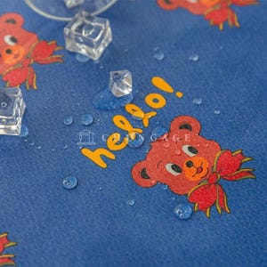 Pode incluir: Tecido azul com um padrão repetido de ursinhos vermelhos com laços e a palavra "hello" em amarelo. Gotas de água e cubos de gelo estão espalhados pela superfície.