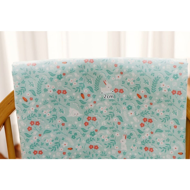 May include: A light blue fabric with a repeating pattern of white bunnies, small red flowers, and green leaves. The fabric is 2 cm wide.
