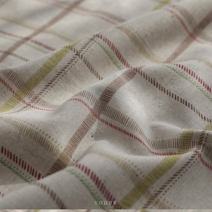 Puede incluir: Primer plano de una tela beige con un patrón de cuadros. El diseño de cuadros presenta líneas en tonos marrón, rojo y verde. La tela está ligeramente arrugada, creando sombras y profundidad. La textura parece suave y natural.