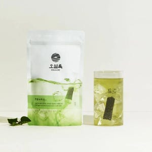 Könnte beinhalten: Ein weißer Beutel mit einem schwarzen Logo, auf dem "OSULLOC" und ein Sonnenbild zu sehen sind. Der Beutel enthält grüne Teebeutel. Im Vordergrund steht ein Glas mit grünem Eistee mit Eiswürfeln und einem Teebeutel.