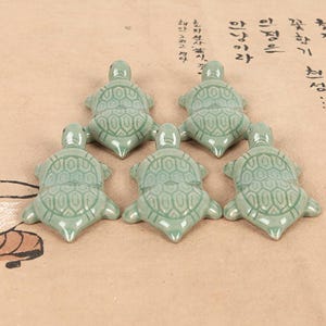 Puede incluir: Cinco figuritas de tortugas de cerámica verde claro dispuestas sobre una superficie marrón claro. Cada tortuga tiene un patrón de caparazón detallado y mira en diferentes direcciones. Las figuritas son pequeñas y parecen ser objetos decorativos.