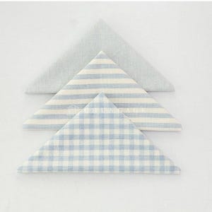 May include: Three folded cloth napkins in a triangle shape. One is solid light blue, one has blue and white stripes, and the other has a blue and white gingham pattern. The napkins are arranged on a white surface.