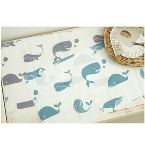 以下が含まれることがあります： 白地に青とグレーのクジラの繰り返し模様が描かれた布。クジラは泳いだり、ジャンプしたり、泡を吹いたりするなど、さまざまなポーズをとっています。