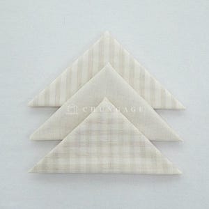 May include: Three folded cloth napkins in a triangular shape. The napkins have different patterns: vertical stripes, a solid color, and a plaid design. The colors are neutral, with a beige and off-white palette.