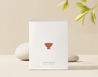 May include: A white box of Cinnamon Ginger Tea with a minimalist design. The box features a small, reddish-brown bowl icon and text in Korean and English. The tea box is presented on a neutral background with a few decorative elements.