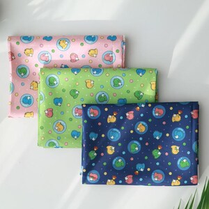 May include: Three folded fabric squares in pink, green, and navy blue, each patterned with cartoon animals in bubbles. The pink square has a light pink background, the green has a light green background, and the navy blue has a dark blue background.