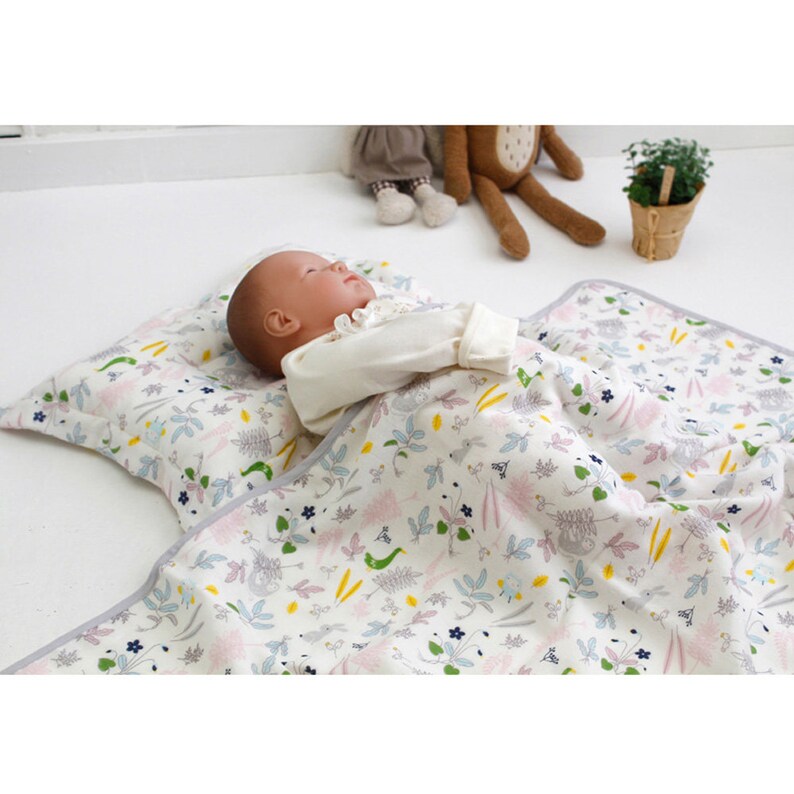 May include: A white baby blanket with a floral pattern and a gray border. A baby doll is lying on the blanket, which is folded in half to create a pillow.