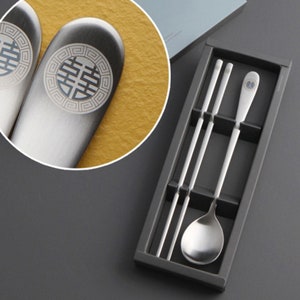 May include: A set of silver chopsticks and a spoon in a black box. The chopsticks and spoon have a decorative design with a Chinese character on them.