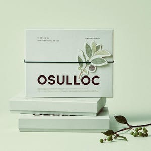 May include: A stack of three light green boxes with the word "OSULLOC" in dark red letters. The top box has a green elastic band and a sticker of a plant with green leaves. The text "TEA VARIATION 30" is printed on the top box.