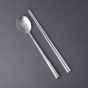May include: A set of silver chopsticks and a spoon. The chopsticks are laid out parallel to each other, while the spoon is placed diagonally across them.