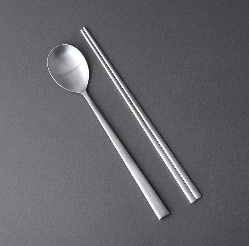 May include: A set of silver chopsticks and a spoon. The chopsticks are laid out parallel to each other, while the spoon is placed diagonally across them.