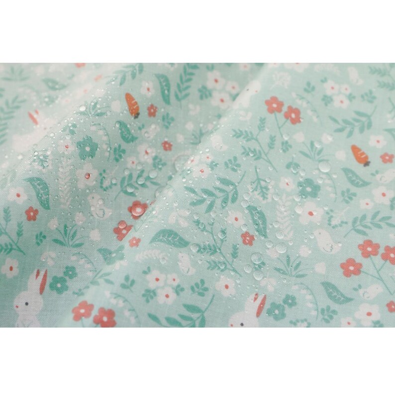May include: A light green fabric with a white bunny rabbit and floral pattern. The fabric is covered in small water droplets.
