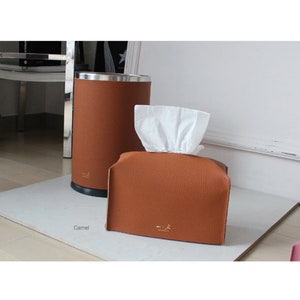 Puede incluir: Una caja de pañuelos de cuero marrón con una caja de pañuelos de papel blanca en su interior. La caja de pañuelos de cuero tiene un logotipo dorado que dice "Bespoke". La caja de pañuelos está sobre una superficie blanca. La palabra "Camel" está escrita en la superficie.