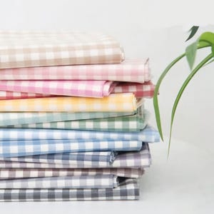 Könnte beinhalten: Ein Stapel Gingham-Stoff in verschiedenen Farben, darunter Beige, Rosa, Gelb, Blau und Grau. Der Stoff ist ordentlich gefaltet und zu einem Stapel gestapelt.