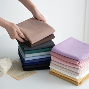 以下が含まれることがあります： 茶色、グレー、緑、青、紫など、さまざまな色の折り畳まれた布製ナプキンの山。 ナプキンはきちんと配置されており、手に茶色のナプキンが上に持たれています。 ピンクと黄色のナプキンの2番目のスタックも表示されます。