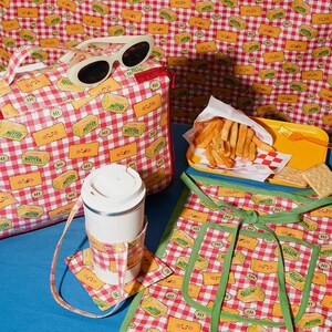 May include: A collection of picnic-themed accessories, including a tote bag, sunglasses, a travel mug with a coaster, and an apron. The items feature a red and white checkered pattern with smiling butter stick graphics. A yellow tray of fries and crackers are also present.