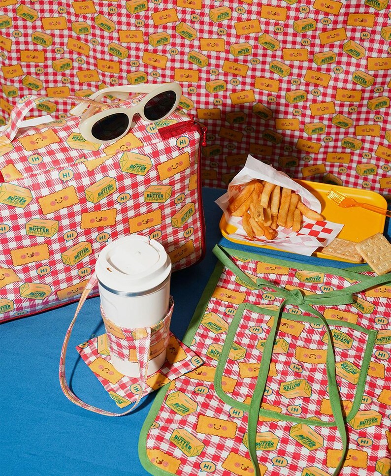 May include: A collection of picnic-themed accessories, including a tote bag, sunglasses, a travel mug with a coaster, and an apron. The items feature a red and white checkered pattern with smiling butter stick graphics. A yellow tray of fries and crackers are also present.