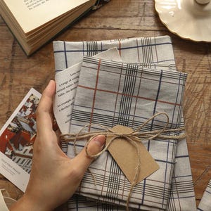 Puede incluir: Un conjunto de servilletas de tela a cuadros beige y gris, atadas con hilo y una etiqueta en blanco. Las servilletas están dobladas, lo que sugiere un juego. Un libro antiguo y una tarjeta pequeña están en el fondo.