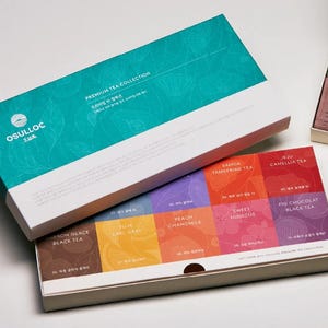 Op de afbeelding: Een witte doos met een turquoise deksel waarop "OSULLOC" en "PREMIUM TEA COLLECTION" staat. De doos is open en laat een set van tien theezakjes zien, elk met een andere smaak en ontwerp. De smaken zijn Marron Glace Zwarte Thee, Yuja Earl Grey, Perzik-Kamille, Samda Mandarijn Thee, Zoete Hibiscus, Jeju Kamelia Thee en Vijg-Chocolade Zwarte Thee.