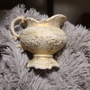 Könnte beinhalten: Eine weiße Keramikkanne mit goldenen Akzenten und einem floralen Design. Die Kanne hat einen Henkel und einen breiten Boden.