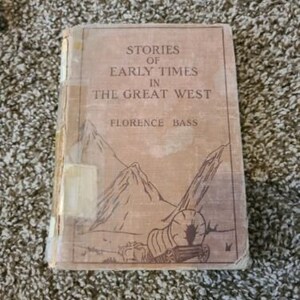 May include: A vintage book titled "Stories of Early Times in the Great West" by Florence Bass. The cover features a brown background with a drawing of a covered wagon and mountains.