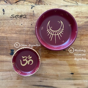 May include: Set of two red ceramic incense holders with gold painted designs. One holder features the Om symbol and the other has a crescent moon with rays of light. The text "Set #2" is written on the wooden surface.