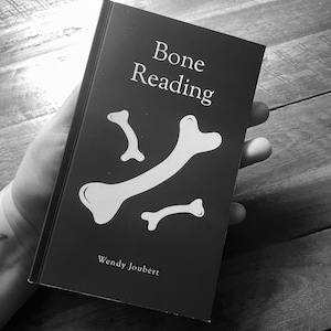 May include: A black and white book titled "Bone Reading" with white bone illustrations. The author's name, Wendy Joubert, is printed at the bottom. The book is held in a hand against a wooden surface.