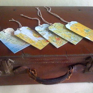 May include: Five rectangular luggage tags with map designs are arranged on a brown suitcase. Each tag displays a different map section, with string attached. The suitcase has a handle and metal clasps.