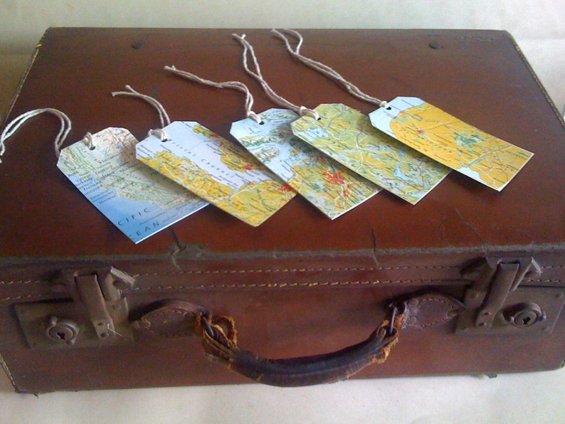 May include: Five rectangular luggage tags with map designs rest on a brown suitcase. Each tag features a different section of a map, with a string attached for tying. The suitcase has a handle and metal clasps.