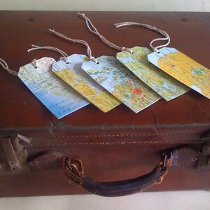 May include: Five rectangular luggage tags with map designs and twine strings rest on a vintage brown suitcase. The tags feature various map sections in shades of blue, yellow, and green, suggesting a travel theme. The suitcase has a leather handle and metal clasps.