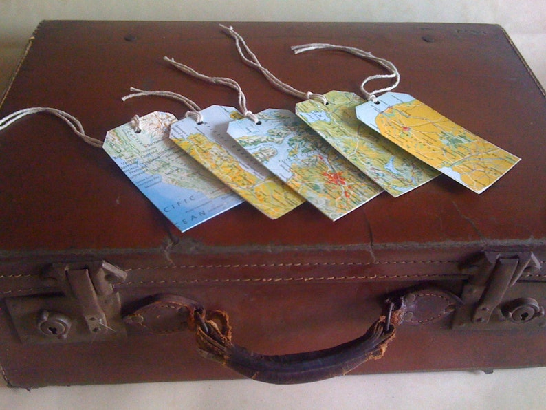 May include: Five rectangular luggage tags with map designs and twine strings rest on a vintage brown suitcase. The tags feature various map sections in shades of blue, yellow, and green, suggesting a travel theme. The suitcase has a leather handle and metal clasps.