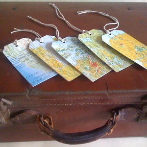 May include: Five rectangular luggage tags with map designs and twine strings rest on a brown suitcase. The tags feature various geographical areas, including the Pacific Ocean. The suitcase has a leather handle and metal clasps.