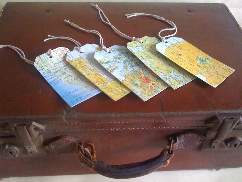 May include: Five rectangular luggage tags with map designs and twine strings rest on a brown suitcase. The tags feature various geographical areas, including the Pacific Ocean. The suitcase has a leather handle and metal clasps.
