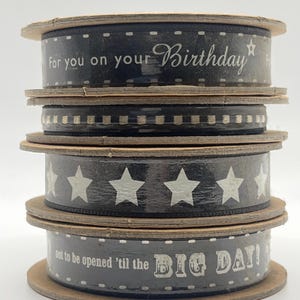 Puede incluir: Una pila de carretes de cinta negra con texto blanco y diseños de estrellas. El texto incluye "For you on your Birthday" y "not to be opened 'til the BIG DAY!". Los carretes tienen bordes de madera y están apilados.