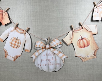 May include: A baby shower banner featuring baby clothes and a pumpkin-shaped decoration. The baby clothes are in the shape of onesies, with pumpkin graphics. The pumpkin decoration has a bow and the text "a little pumpkin is on the way".