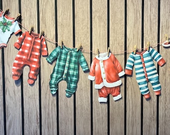 May include: A decorative garland featuring paper cutouts of baby clothing hanging from a string with small wooden clips. The items include a holly-patterned shirt, red plaid sleeper, green plaid sleeper, Santa suit, and blue and white striped sleeper.