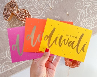 May include: Three rectangular greeting cards in bright pink, orange, and yellow, each featuring the word "diwali" in gold script lettering. The yellow card also includes the text "HAPPY" and a small gold diya lamp icon above the word "diwali".
