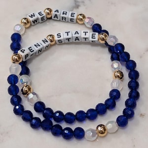 May include: A blue and white beaded bracelet with gold accents. The beads spell out "We Are Penn State".