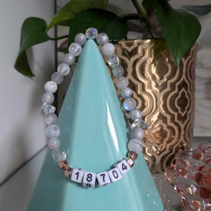 May include: A beaded bracelet with a mix of gray, clear, and iridescent beads. The bracelet includes white letter beads spelling out the numbers "18704". The bracelet is displayed on a light blue pyramid-shaped stand, with a gold decorative pot and plant in the background.