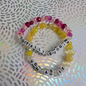 May include: Three beaded bracelets with the phrases "TOTALLY BUGGIN" and "UGH AS IF" spelled out in white letter beads. The bracelets feature a mix of pink, yellow, and clear beads, with a shimmering background.