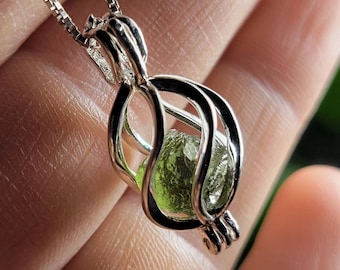 May include: A silver pendant necklace featuring a cage-like design holding a rough, green gemstone. The pendant hangs from a delicate chain. The gemstone has a natural, irregular shape and a vibrant green color.