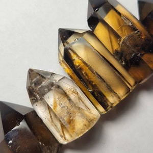May include: Close-up of several smoky quartz crystal points. The crystals have a translucent, amber-brown color with sharp, defined edges. The crystals are arranged diagonally, showcasing their natural formations and textures.
