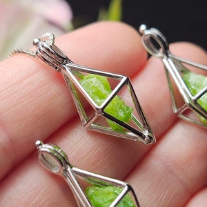 May include: Three silver-colored geometric pendants, each containing a vibrant green gemstone. The pendants are diamond-shaped cages with a loop at the top for a chain. The gemstones have a rough, natural texture. The background is blurred with hints of pink and purple.