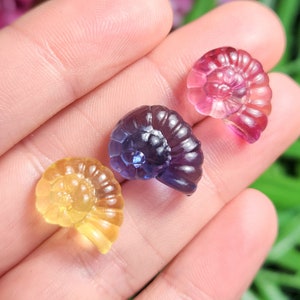 Peut inclure: Trois petites sculptures de pierres pr&eacute;cieuses translucides en forme de spirale, dans les tons jaune, violet et rose. Les sculptures sont tenues dans une main, avec un arri&egrave;re-plan flou de fleurs roses et de feuillage vert.