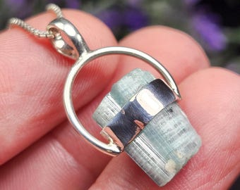 May include: A silver pendant necklace featuring a raw, light blue-green crystal. The crystal is held in place by a silver band and a circular silver bail, suspended from a delicate silver chain. The pendant is a unique piece of jewelry.