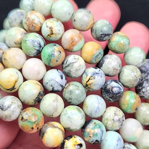May include: A strand of round gemstone beads in various colors, including green, yellow, and cream. The beads have a marbled appearance with unique patterns and textures. The beads are held in a hand, showcasing their size and detail.