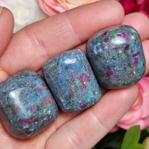 May include: Three tumbled stones of blue kyanite with red ruby inclusions. The stones are smooth and polished, and they are arranged in a row in a hand.