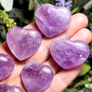 May include: Six heart-shaped amethyst crystals in various sizes, displayed on a hand. The crystals are a deep purple color and have a smooth, polished finish.