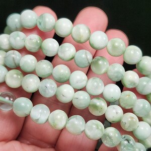 Peut inclure: Plusieurs bracelets de perles vert clair. Les perles sont rondes et ont un aspect marbr&eacute; avec des variations de couleur. Les bracelets sont tenus dans une main sur fond sombre.